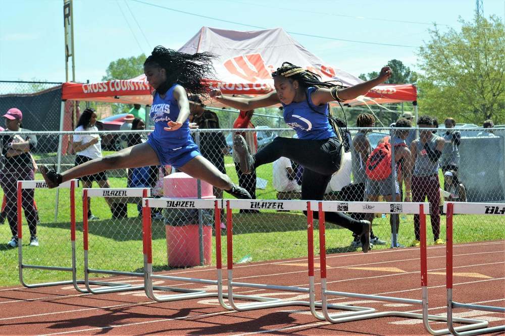 2 students jumping over hurdles during a track meet; wearing blue tanks